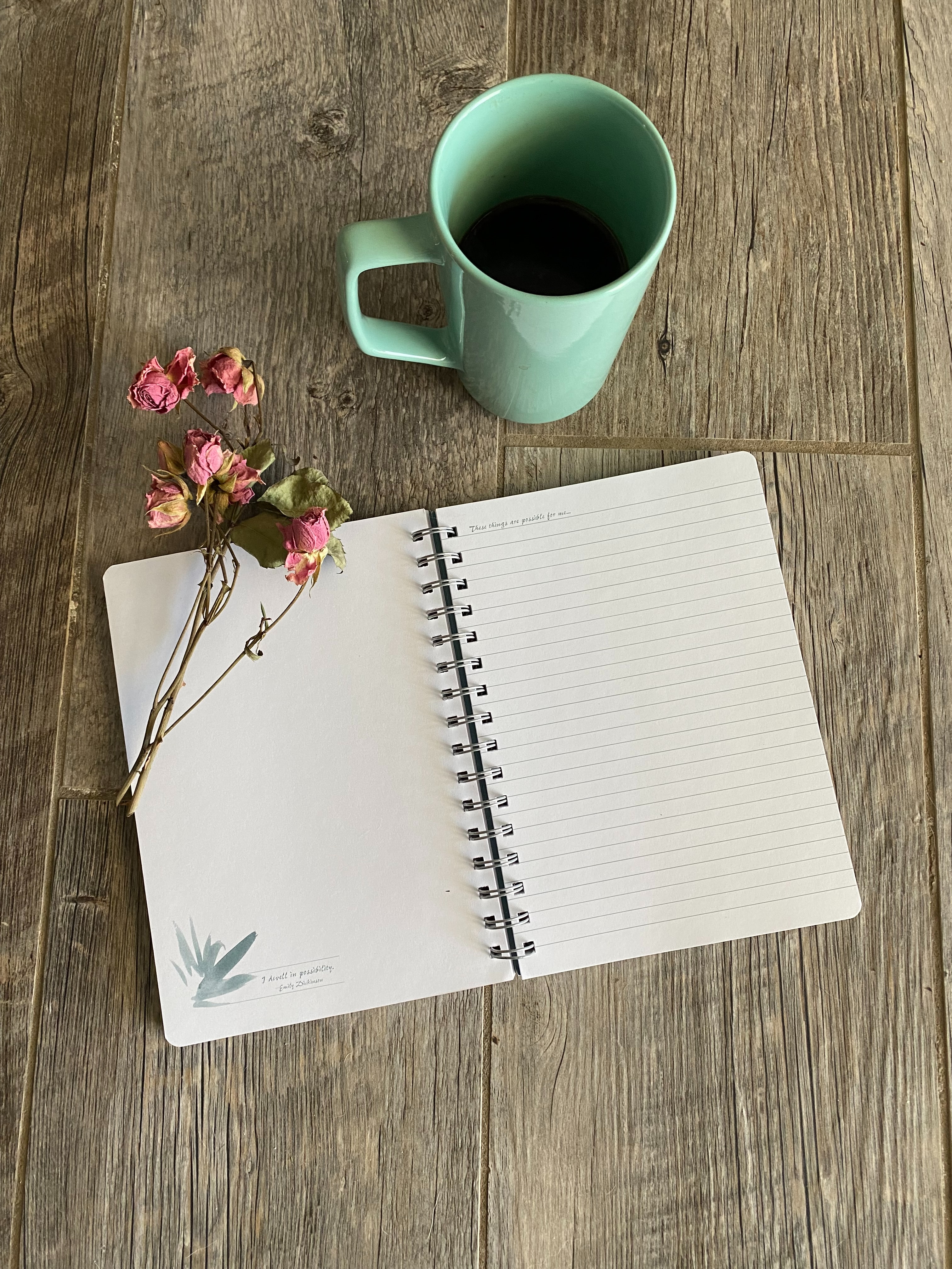Empty page of an open notebook on a wooden table, with a green coffee cup above it. There are some dried roses on the left page of the notebook.